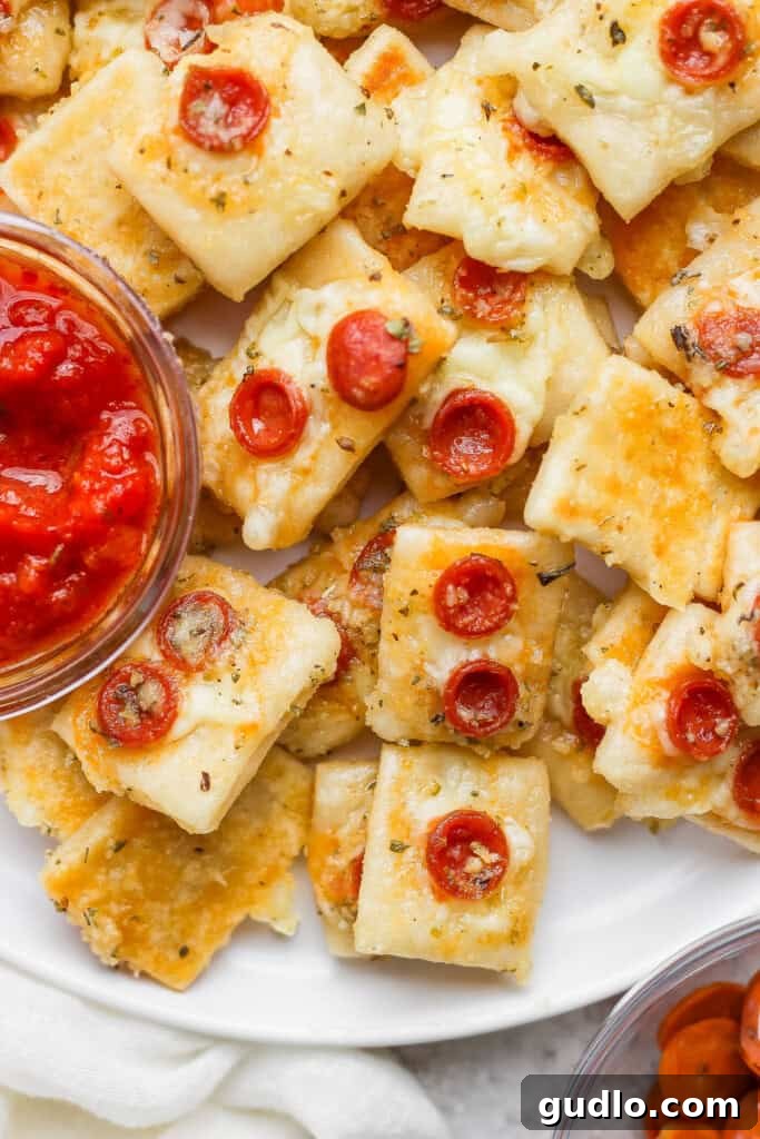 Protein Packed Pepperoni Pizza Bites 12 A plate of square mini pizza bites topped with cheese and small pepperoni slices, served with a bowl of marinara sauce for dipping.