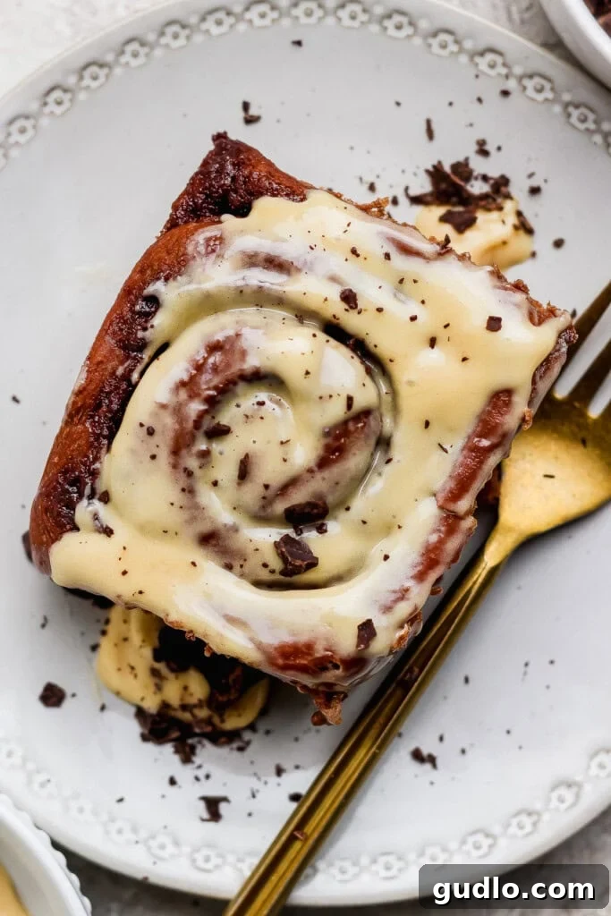 A perfectly baked chocolate cinnamon roll generously topped with a rich cream cheese icing and delicate chocolate shavings, beautifully presented on a pristine white plate with an elegant gold fork beside it.
