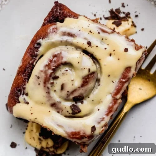 A close-up shot of a single chocolate cinnamon roll, adorned with generous dollops of white cream cheese frosting and sprinkled with fine chocolate shavings, resting on a white plate next to a gold fork, ready to be enjoyed.