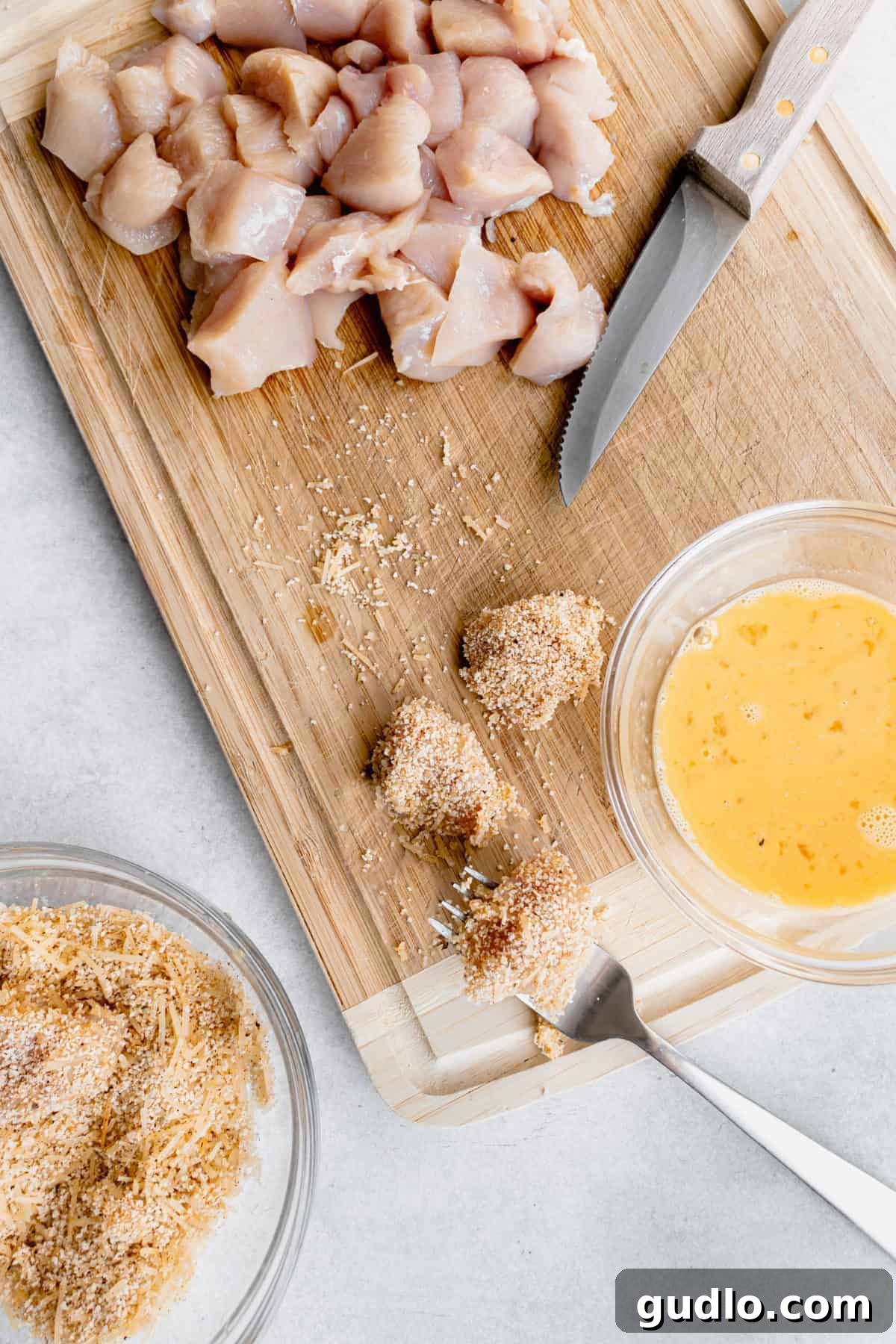 The meticulous coating process for Parmesan Chicken Nuggets. Chicken pieces being dipped into an egg wash and then into a breadcrumb and Parmesan mixture on a cutting board.