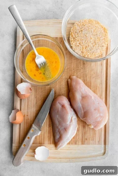 A cutting board with cubed chicken, a bowl of egg wash, and a bowl of seasoned breadcrumb mixture ready for dredging.