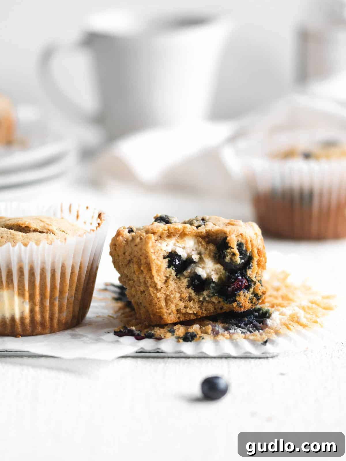 Decadent Blueberry Cream Cheese Muffins with a Hidden Filling A perfectly baked blueberry cream cheese muffin in a delicate paper wrapper, showcasing its golden-brown top, a generous sprinkle of blueberries, and the promise of a creamy filling.