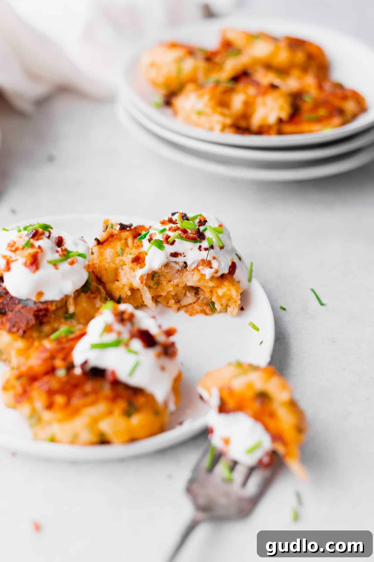 Cheesy Potato Pancakes with Greek Yogurt and Chives Close-up of a stack of cheesy potato pancakes topped with Greek yogurt and finely chopped chives, presented on a white plate.