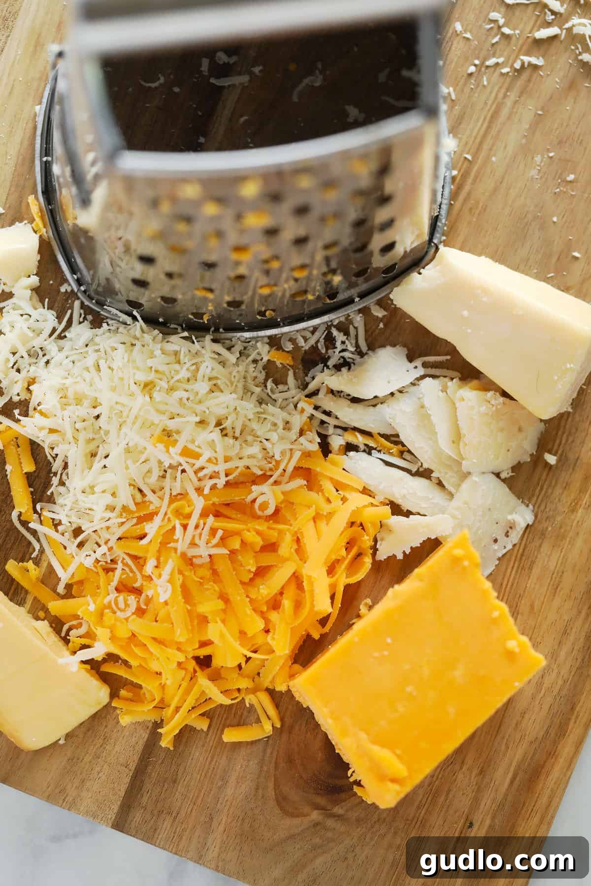 A robust box grater positioned next to piles of various shredded cheeses on a cutting board.