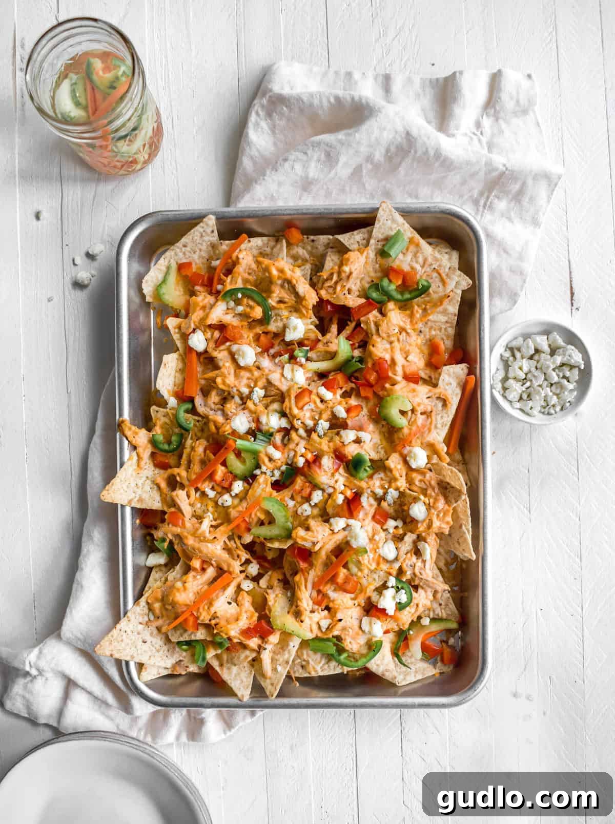 A large baking sheet filled with freshly baked Buffalo Chicken Nachos, piled high with creamy, shredded buffalo chicken, melted cheddar, crumbled blue cheese, and garnished with vibrant quick-pickled carrots, celery, and green onions. It's the ultimate party food.