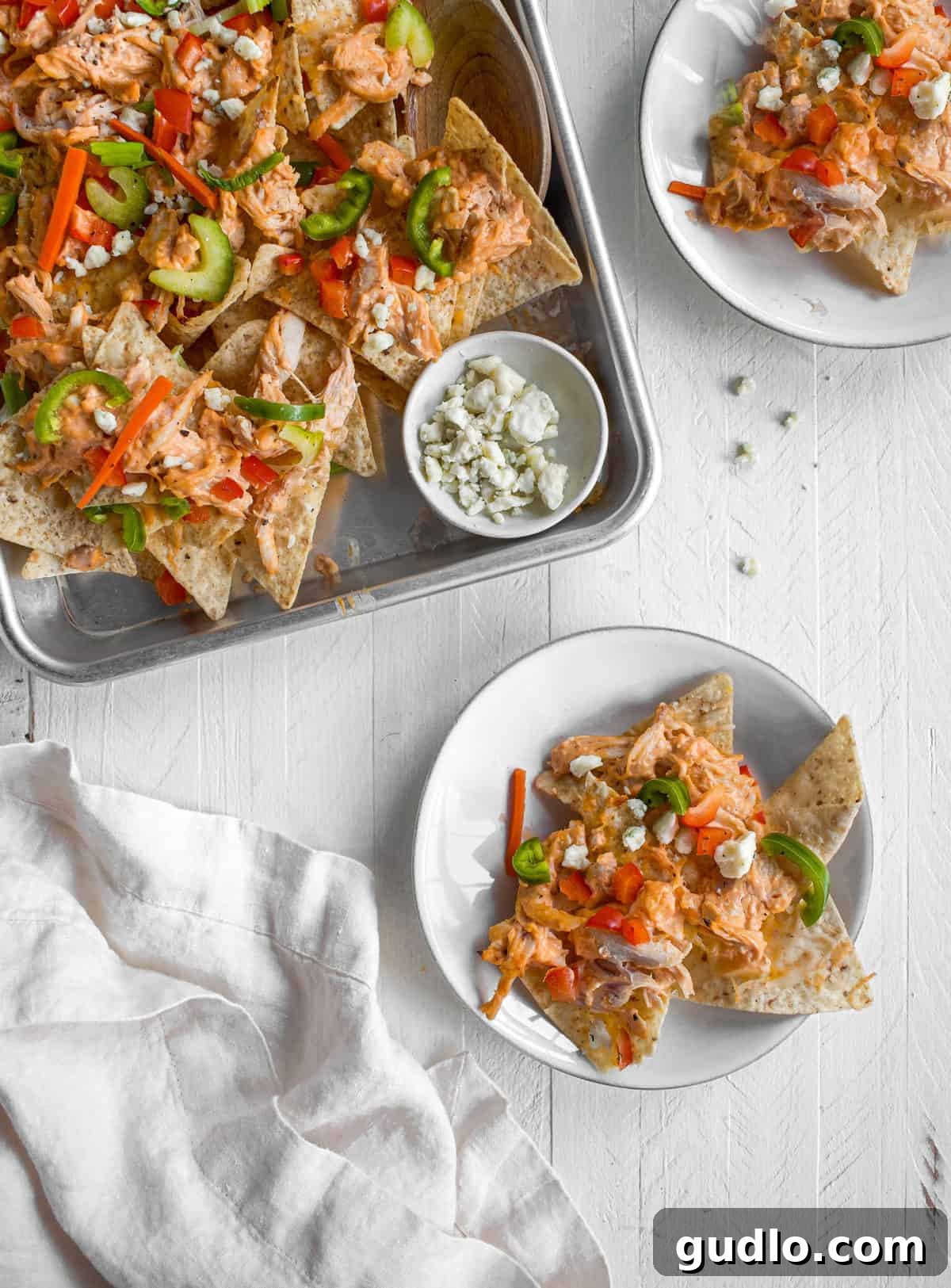 A vibrant display of Buffalo Chicken Nachos served on a rustic baking pan and individual white plates, beautifully garnished with fresh cilantro, a drizzle of ranch dressing, and extra blue cheese crumbles. Perfect for sharing at any festive gathering!