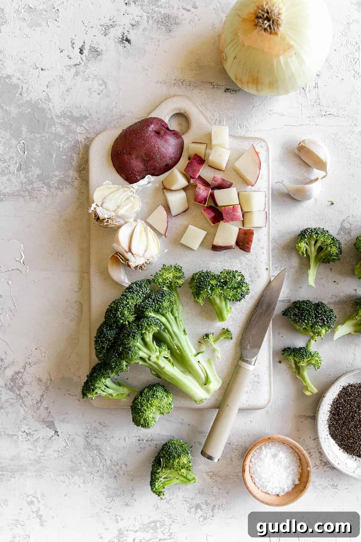 Velvety Broccoli & Cheddar 4 Fresh broccoli, a white onion, a head of garlic, and a red potato laid out on a cutting board, ready for prep.