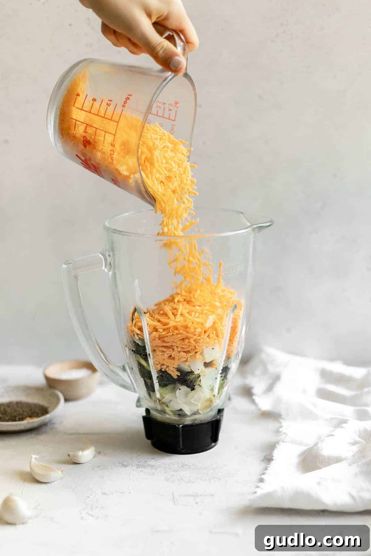 Velvety Broccoli & Cheddar 7 A person adding shredded cheddar cheese into a high-speed blender, creating a vortex of creamy goodness for broccoli cheese soup.
