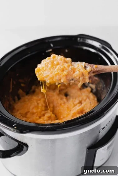 A wooden spoon stirs a crockpot of cheesy macaroni and cheese, showing its creamy texture.