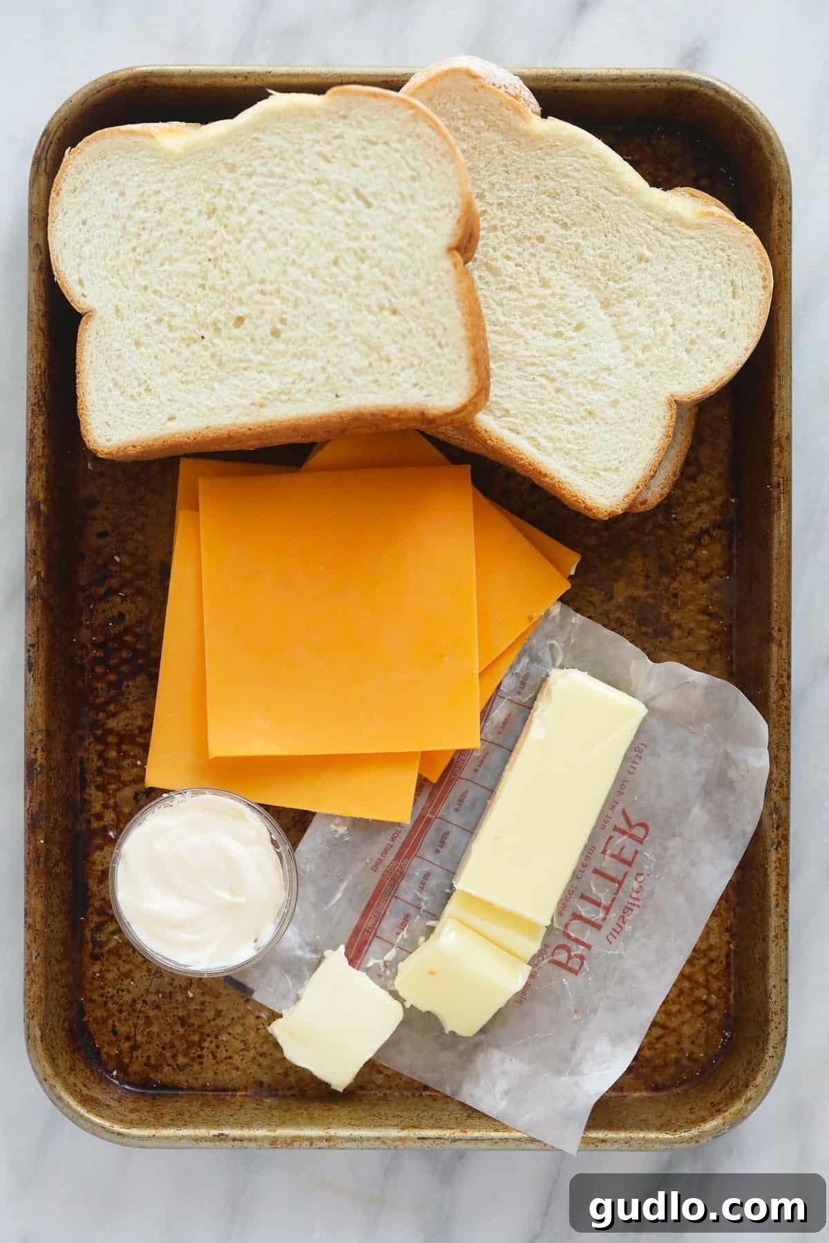 Gather your ingredients for the best sheet pan grilled cheese. Fresh ingredients for sheet pan grilled cheese including slices of sourdough bread, cheddar cheese, a stick of butter, and a jar of mayonnaise, laid out on a kitchen counter.