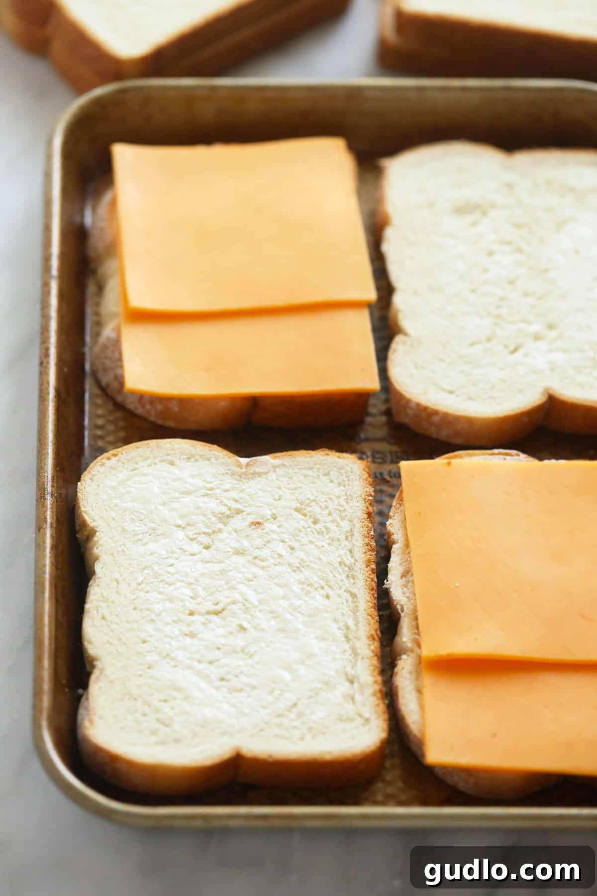 Assembling sheet pan grilled cheese sandwiches for baking. Sheet pan filled with unbaked grilled cheese sandwiches, carefully assembled with buttered bread and cheese layers, ready to be placed in the oven.