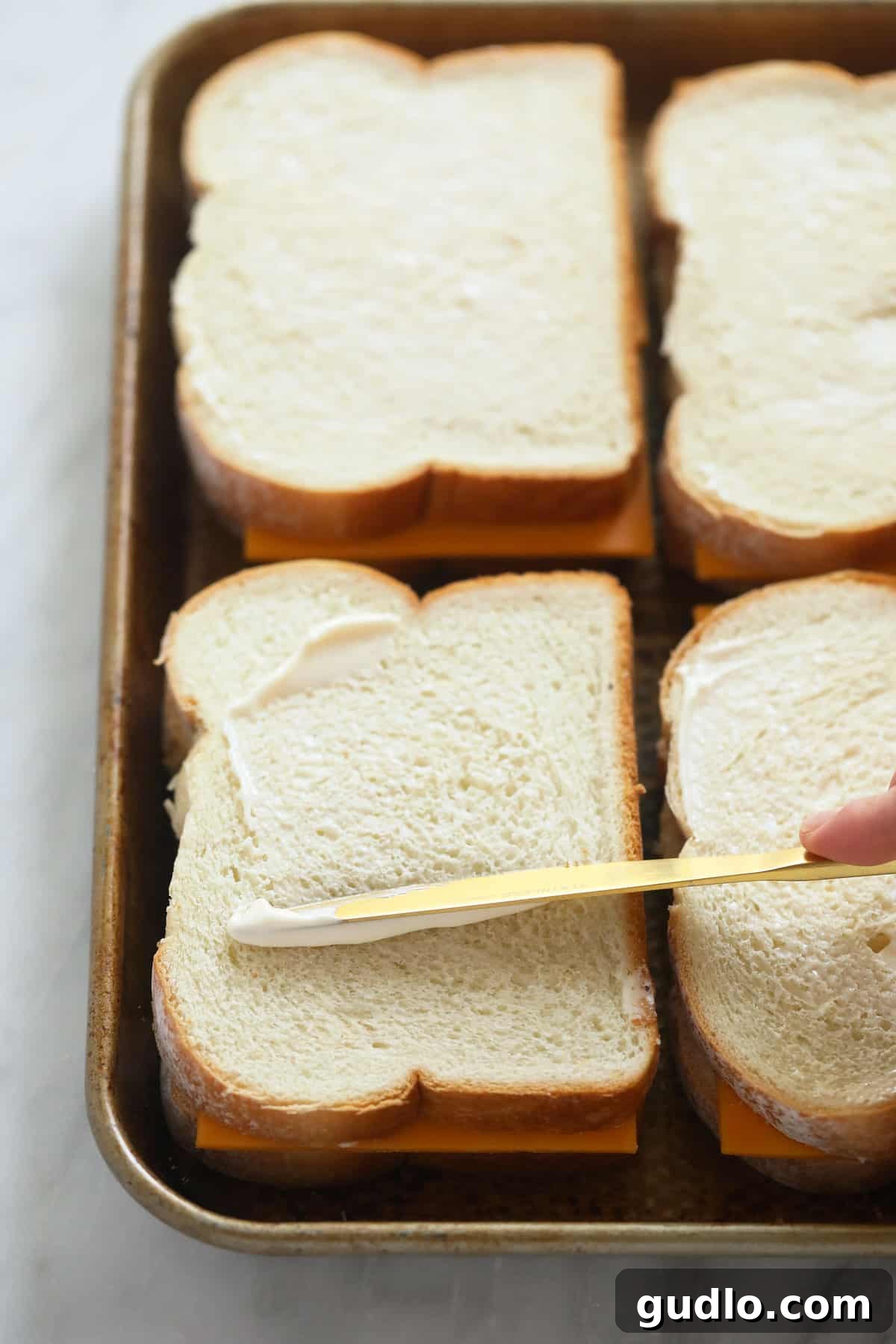 Buttering the bread for sheet pan grilled cheese. A hand gently spreading softened butter onto the outside of a slice of bread on a sheet pan, preparing it for the ultimate sheet pan grilled cheese.