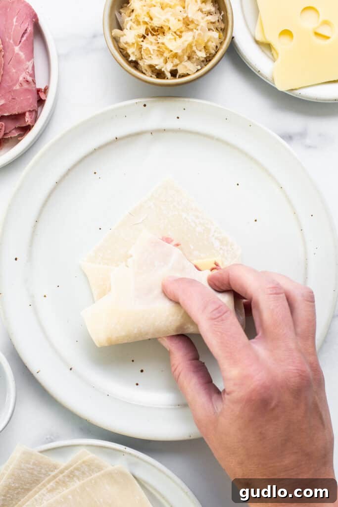 Hand placing a slice of Swiss cheese onto an egg roll wrapper with other ingredients nearby.