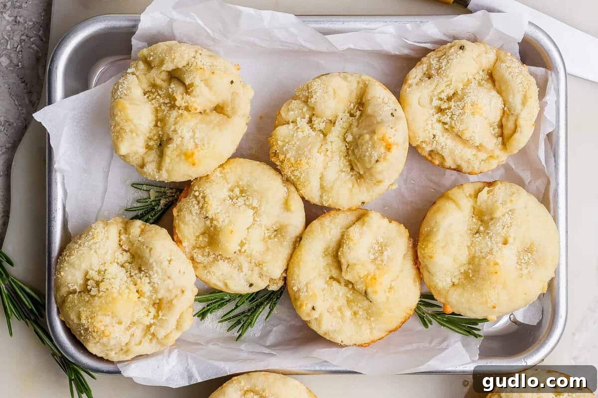 Seven round, golden-brown bread rolls topped with grated cheese are arranged on a parchment-lined metal tray, garnished with sprigs of rosemary.
