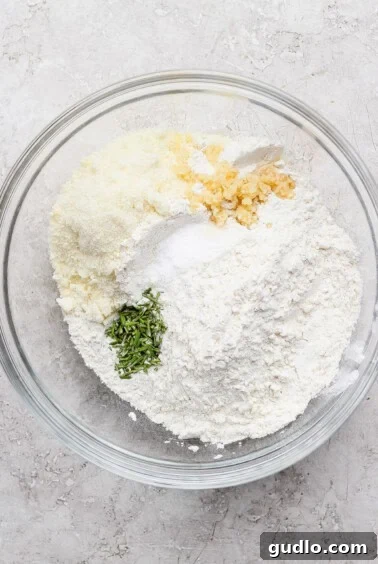 A glass bowl containing flour, grated cheese, chopped garlic, salt, and chopped fresh herbs on a light textured surface.