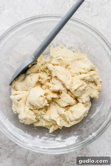 A glass bowl containing a rough, unbaked dough mixture with a spatula resting on the side.
