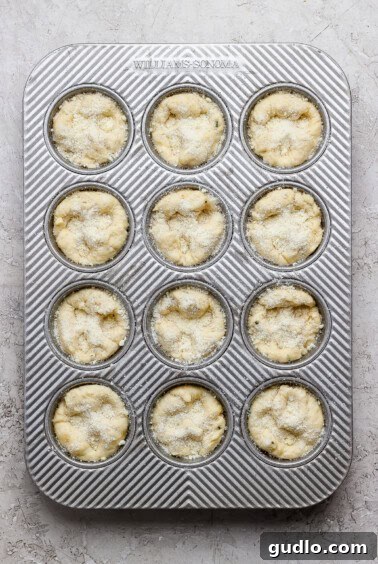A metal muffin tin with twelve unbaked muffin batter portions, sprinkled with sugar, placed on a light textured surface.