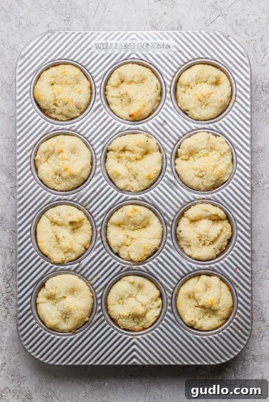 A muffin tin containing twelve baked muffins with a light golden top, set on a textured white surface.