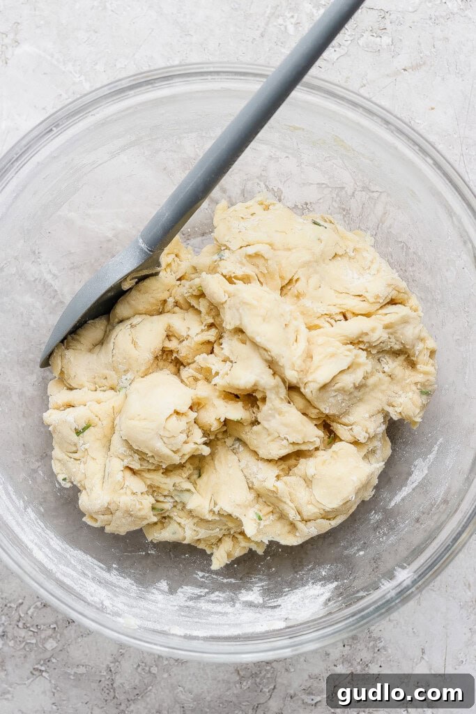 A glass bowl containing a rough, unbaked dough mixture with a spatula resting on the side.