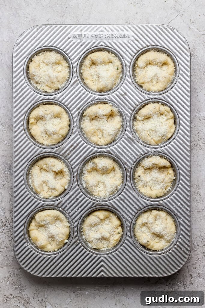 A metal muffin tin with twelve unbaked muffin batter portions, sprinkled with sugar, placed on a light textured surface.
