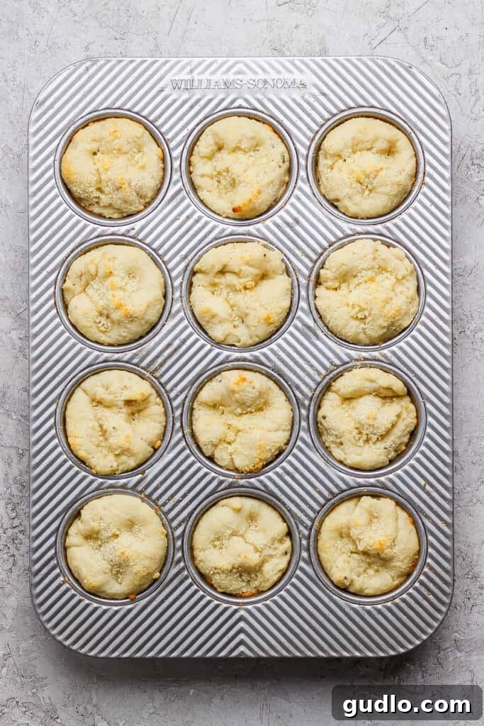 A muffin tin containing twelve baked muffins with a light golden top, set on a textured white surface.