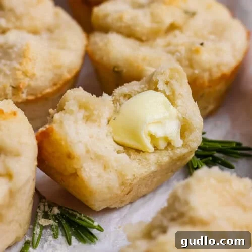 A close-up of a biscuit broken open with a pat of butter melting inside, garnished with fresh rosemary sprigs.