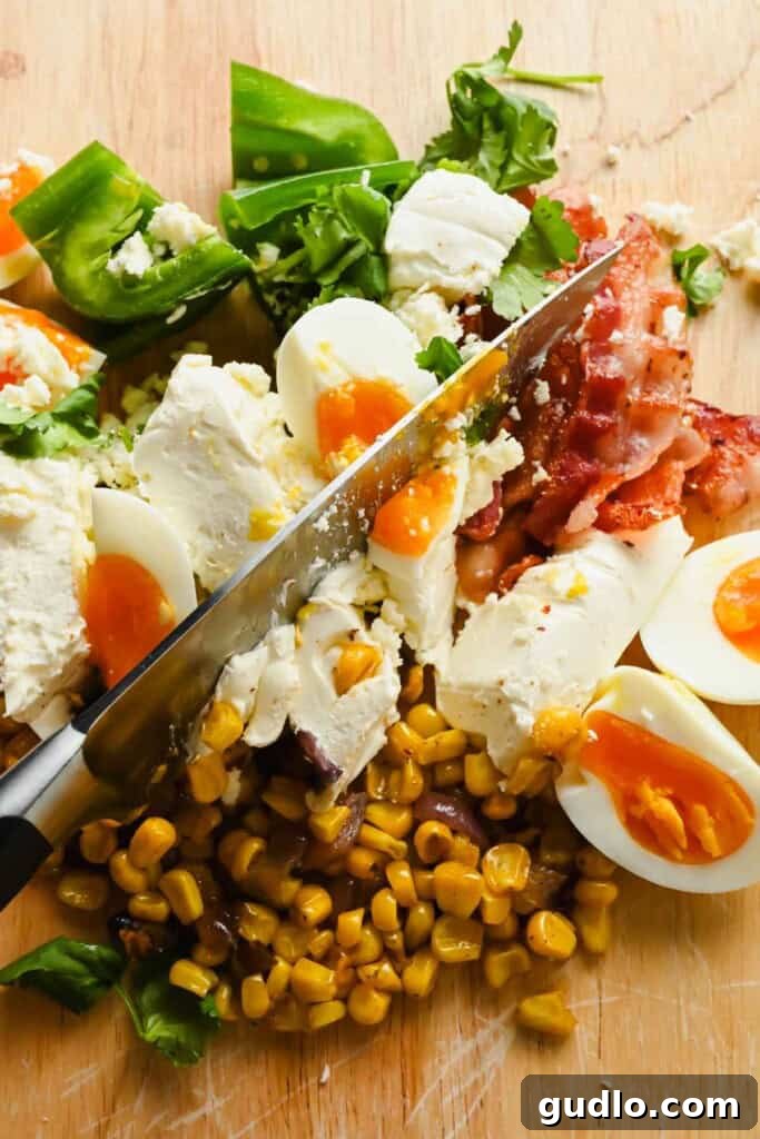 A large chef's knife beginning to chop through the pile of ingredients for the bagel spread on a wooden cutting board.