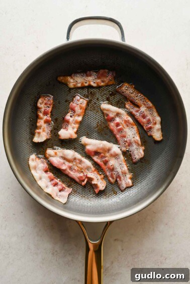 Strips of thick-cut bacon cooking and crisping in a large skillet.