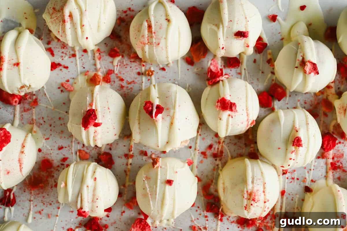 Rows of white chocolate-coated truffles are topped with red crushed pieces and drizzled with white chocolate, arranged on a white surface with scattered crumbs.