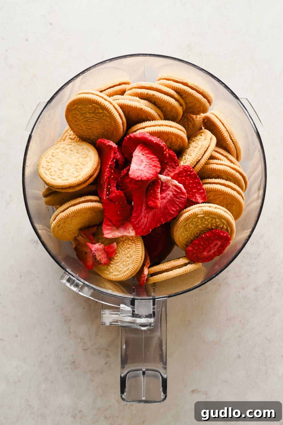 A food processor bowl filled with golden sandwich cookies and freeze-dried strawberry slices on a light surface.