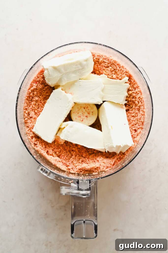 Food processor bowl with crushed pink cookies at the bottom and six blocks of cream cheese on top.