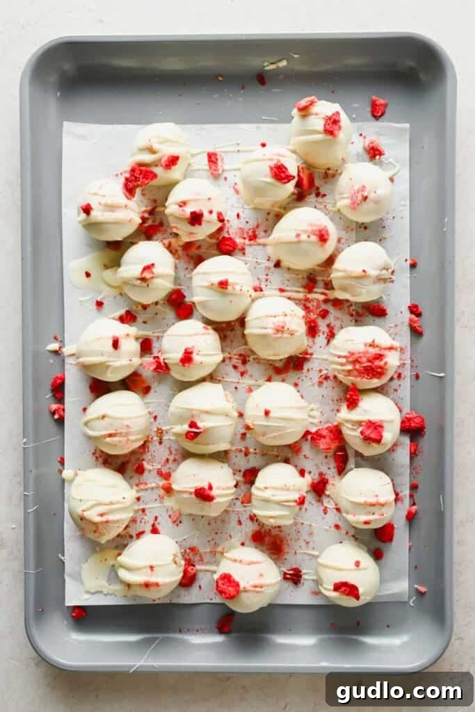 A baking sheet with white chocolate-coated round treats, drizzled with more white chocolate and sprinkled with crushed red pieces, arranged on parchment paper.
