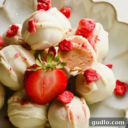 A plate of white chocolate-coated truffles, garnished with freeze-dried strawberry pieces and a halved fresh strawberry. One truffle is cut open to show the filling.