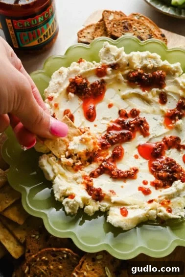 3-Ingredient Boursin Chili Crunch Dip Ready in 5 Minutes 11 A hand dipping a cracker into whipped cheese topped with chili oil, served on a green plate with more crackers on the side.