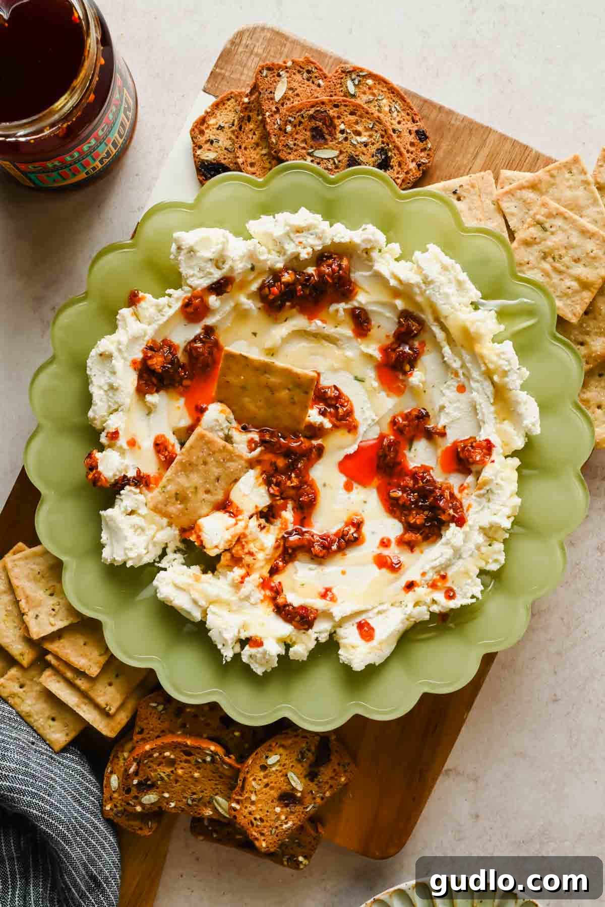 3-Ingredient Boursin Chili Crunch Dip Ready in 5 Minutes 7 Overhead view of boursin chili crunch dip topped with chili oil and honey, served with crackers on a wooden board