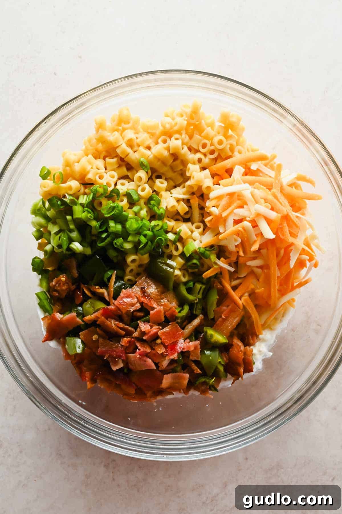 Glass bowl with ditalini pasta, shredded cheddar, chopped green onions, crispy bacon, and roasted jalapeños ready to toss