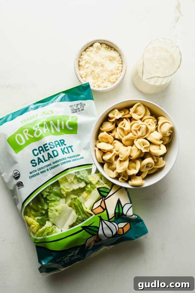 A bag of organic Caesar salad kit, a bowl of grated cheese, a bowl of tortellini pasta, and a small pitcher of creamy dressing on a white surface create the perfect setup for making a delicious tortellini Caesar salad.