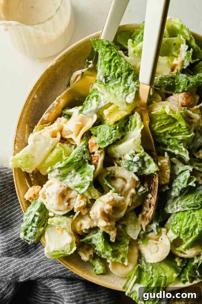 A plate of tortellini Caesar salad with romaine lettuce, croutons, grated cheese, and creamy dressing, served with utensils.