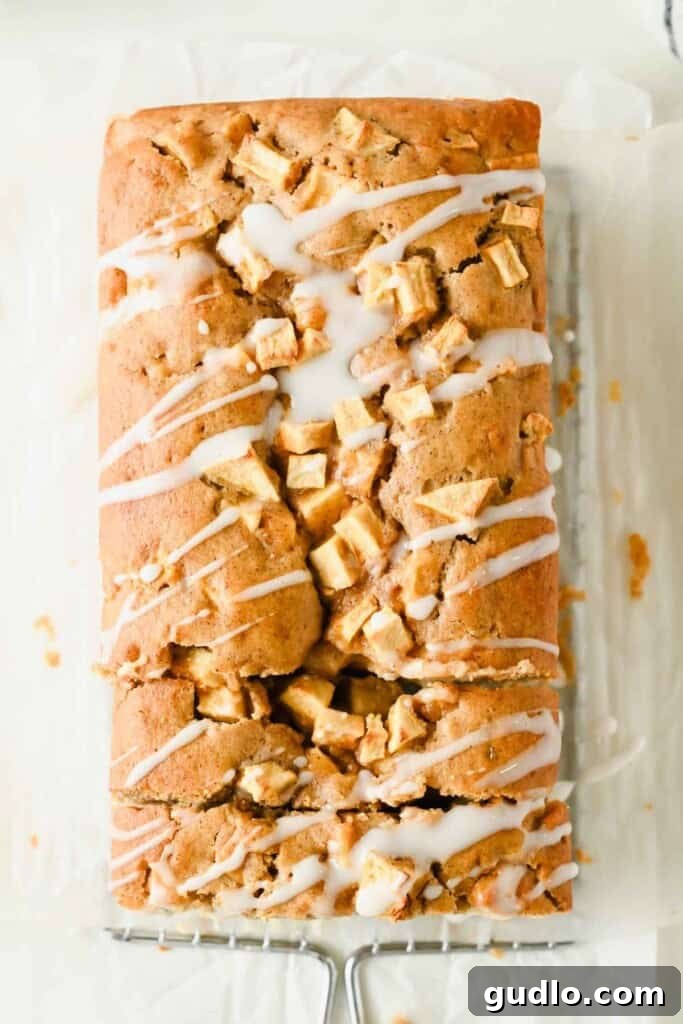 Apple Fritter Bread with Cream Cheese Swirl 9 A loaf of apple bread topped with chopped apple pieces and drizzled with white icing sits on parchment paper and a cooling rack.