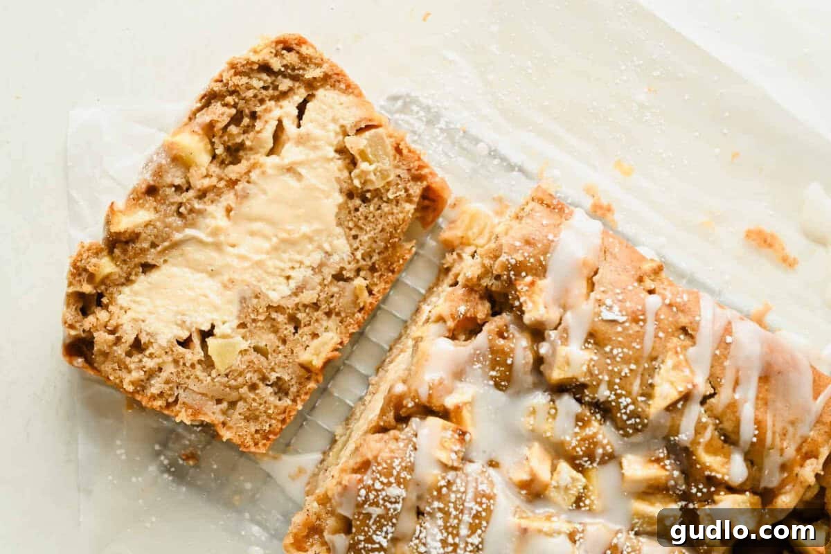 Apple Fritter Bread with Cream Cheese Swirl 10 A sliced loaf of apple bread with visible apple pieces, cream cheese filling, and icing drizzled on top, sitting on parchment paper.