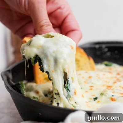A hand lifts a portion of cheesy spinach dip from a skillet, showing melted cheese stretching. Garlic bread slices are visible in the background.